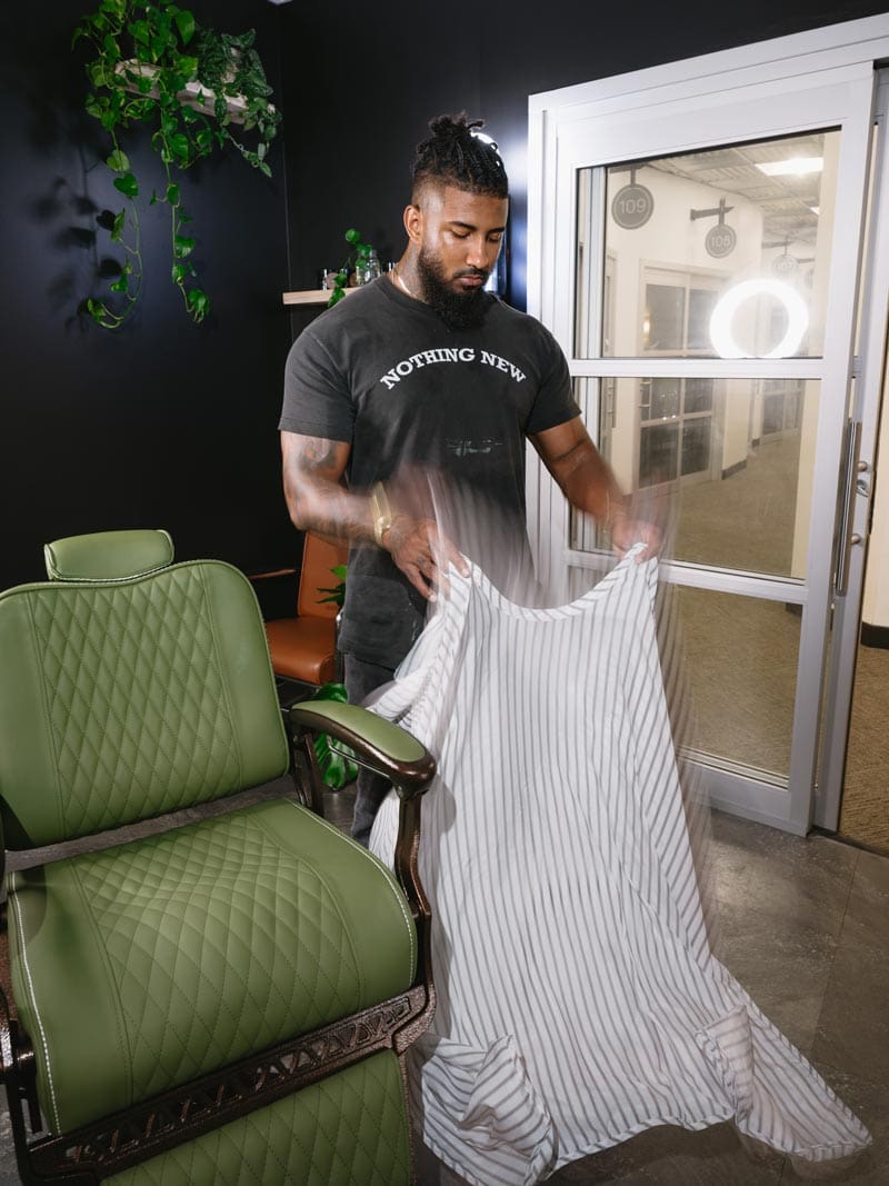 Barber Cipper Cole Knoll preparing a striped robe next to a green chair in a modern Charlotte, NC barbershop studio
