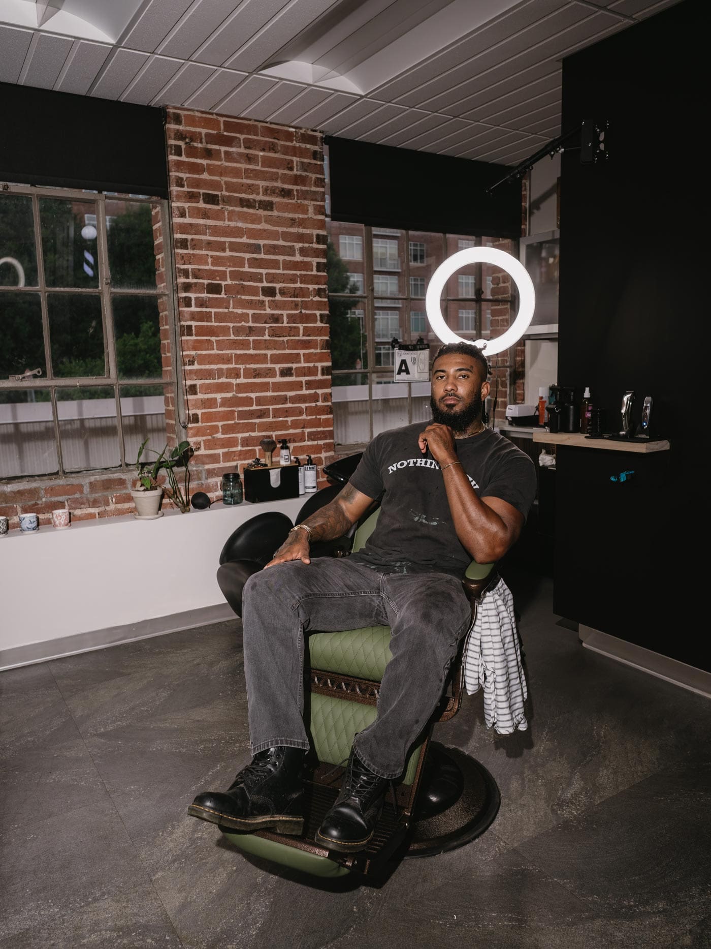 Barber Clipper Cole Knoll seated in a barber chair with brick wall and grooming tools in the background at a Charlotte, NC barbershop